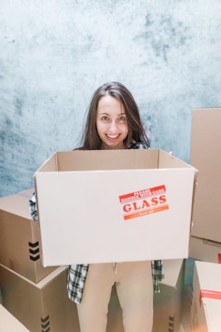A young woman with long brown hair smiling and carrying an empty cardboard box labeled 'Glass' in a room with a textured light blue wall. She is dressed in casual clothing, including a checkered shirt