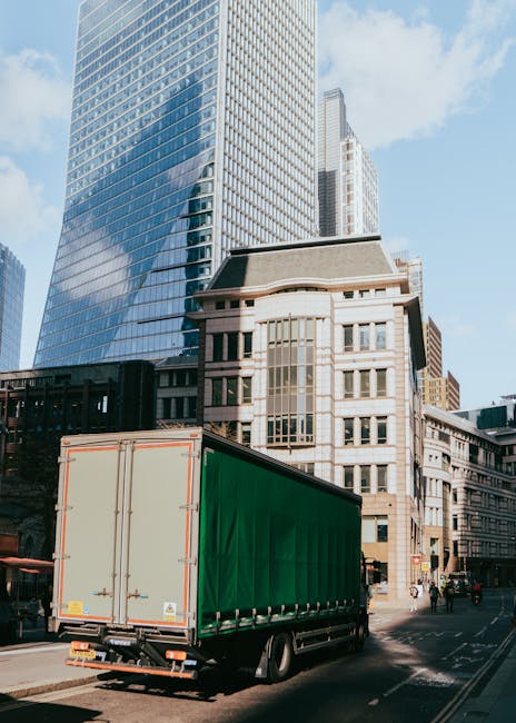 A detailed view of a home relocation scene showing several large cardboard boxes and wrapped furniture being loaded into a white moving van parked outside a residential property. The boxes are stacked